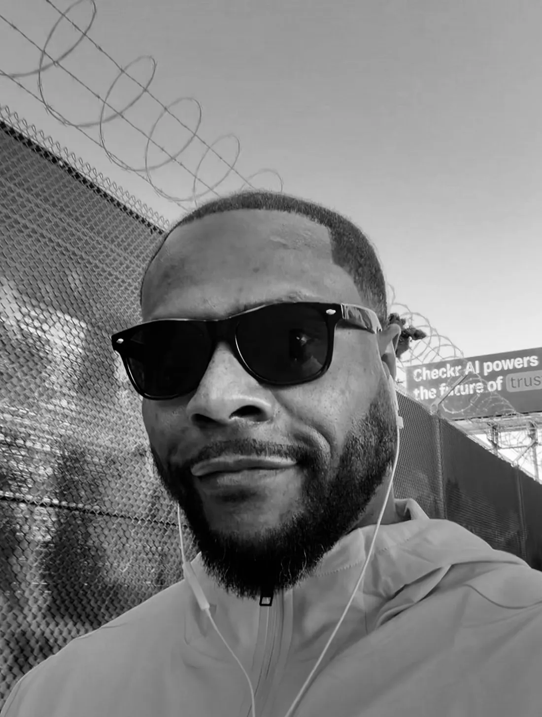 Black-and-white portrait of a man wearing sunglasses, wired earbuds and a zip-up jacket beside a chain-link fence.