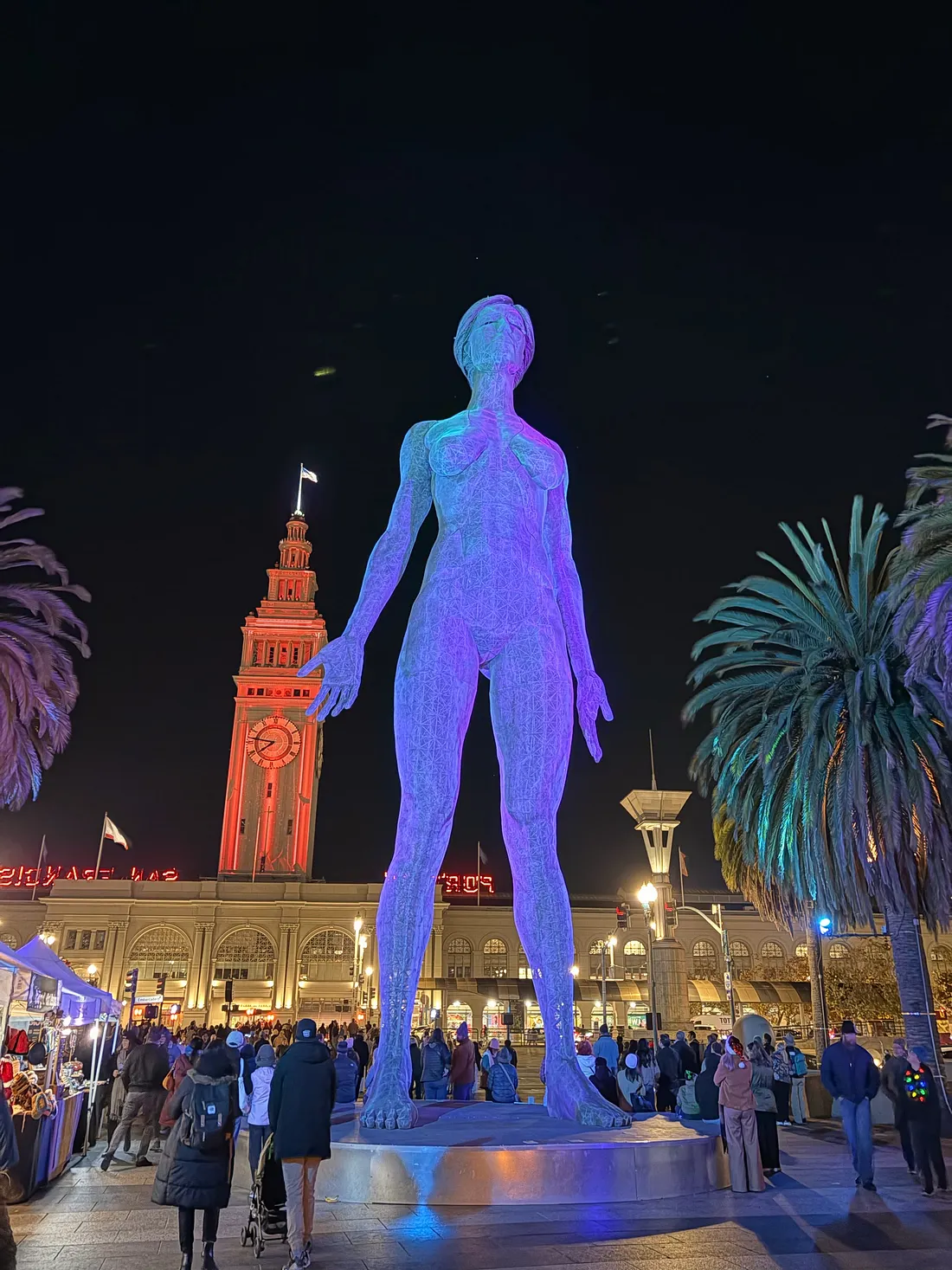 A human-shaped sculpture lit blue and purple in front of the San Francisco Ferry Building clock tower at night.