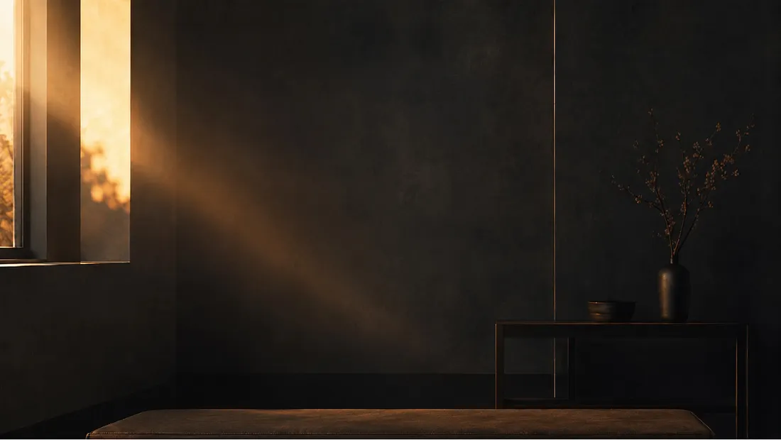 Warm sunlight streams through a window into a dark room with a vase and low table.