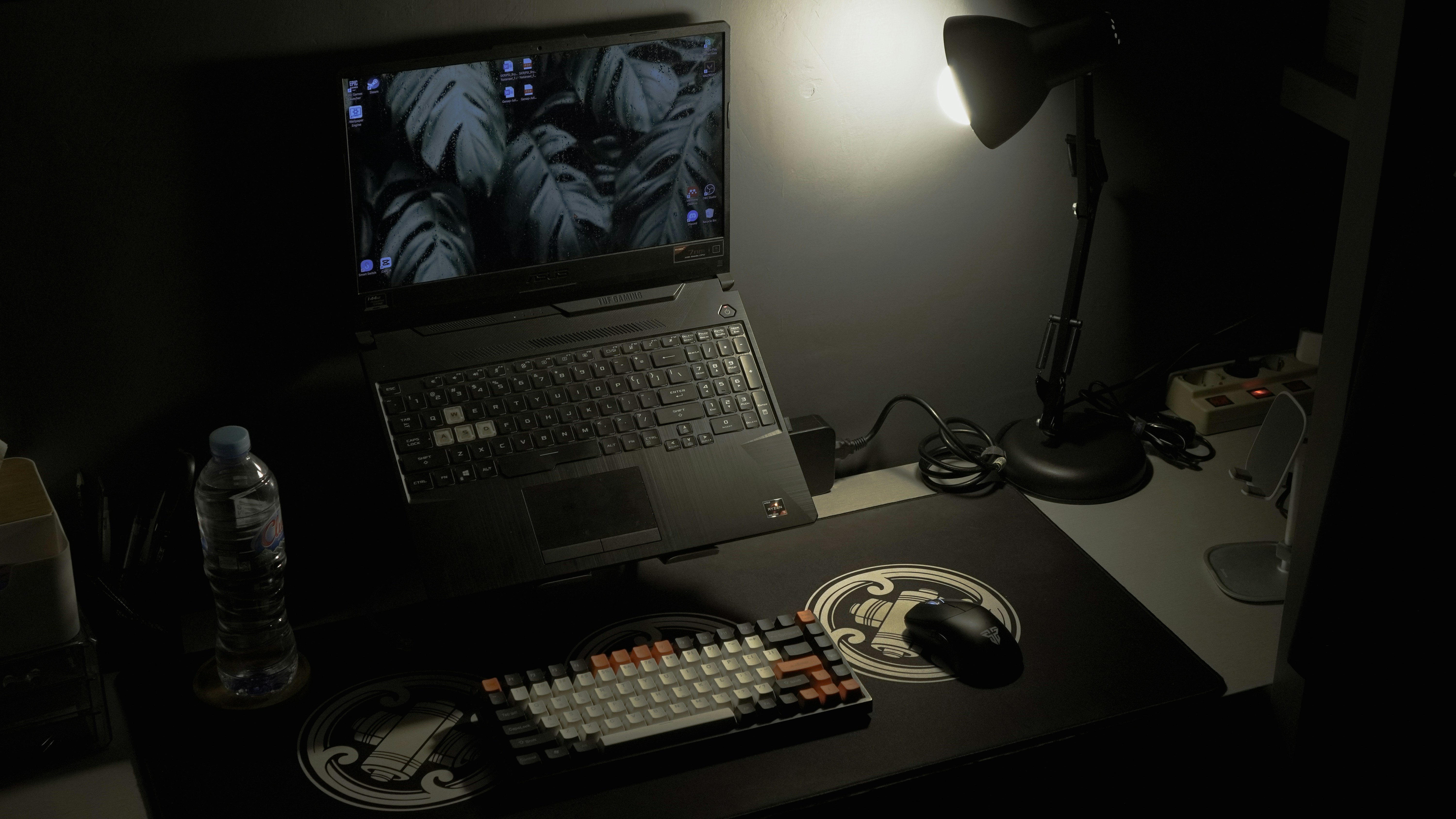A dimly lit desk with an open laptop, desk lamp, keyboard, mouse, and water bottle.