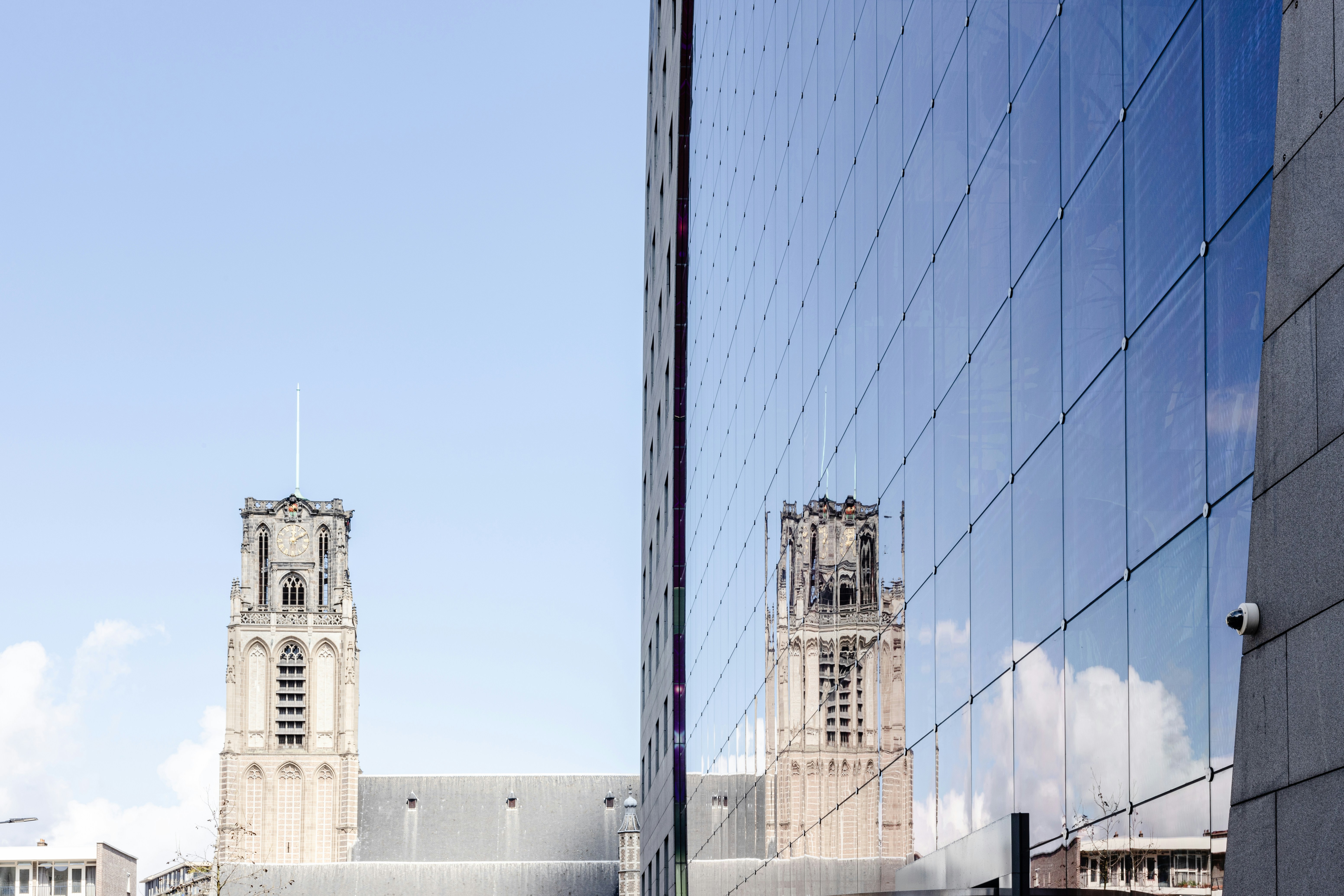 A stone church tower under a blue sky, reflected in the glass facade of a nearby building.