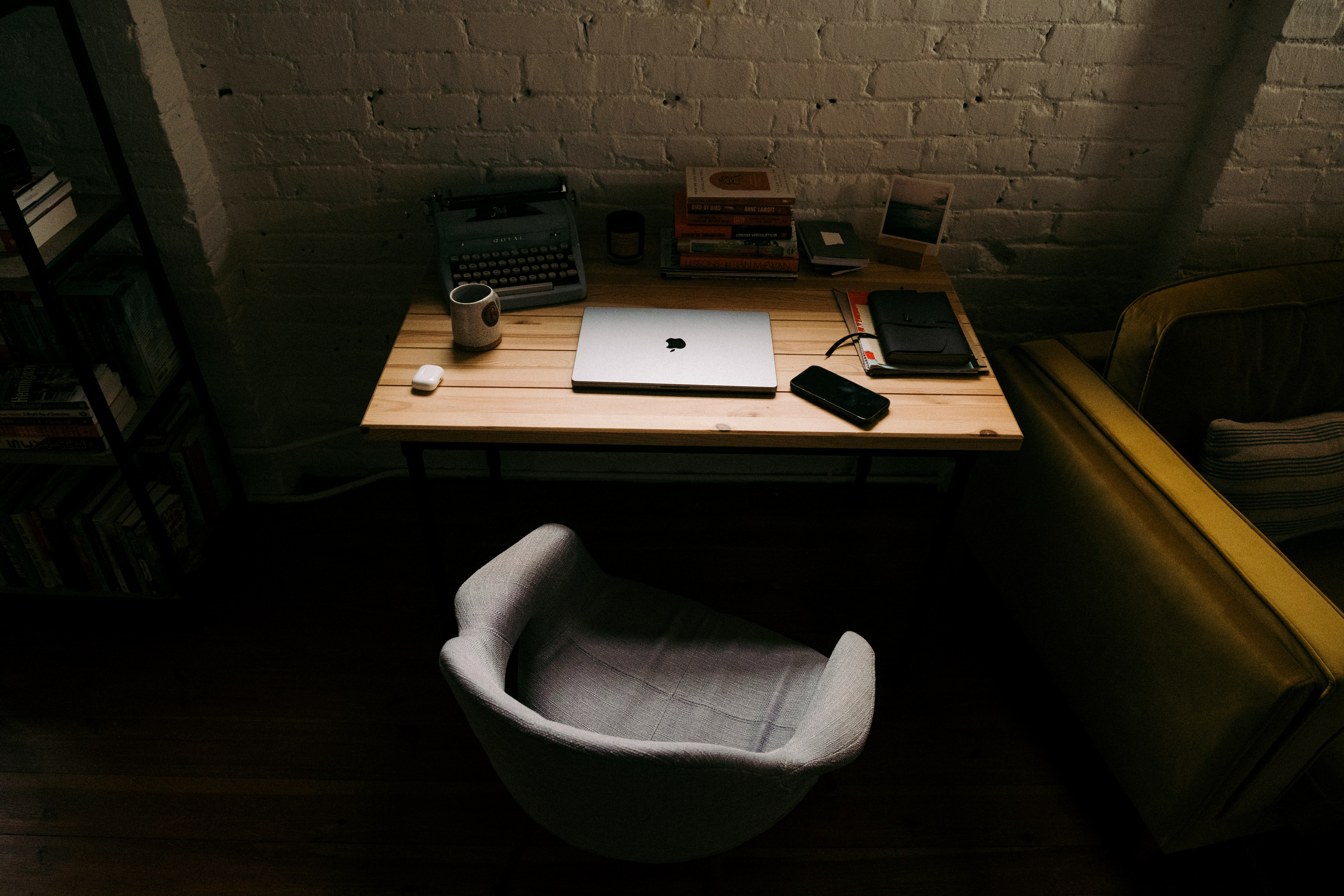 A wooden desk against a white brick wall, with a closed laptop, typewriter, books, notebooks, and a mug, beside a gray chair and mustard-colored sofa.
