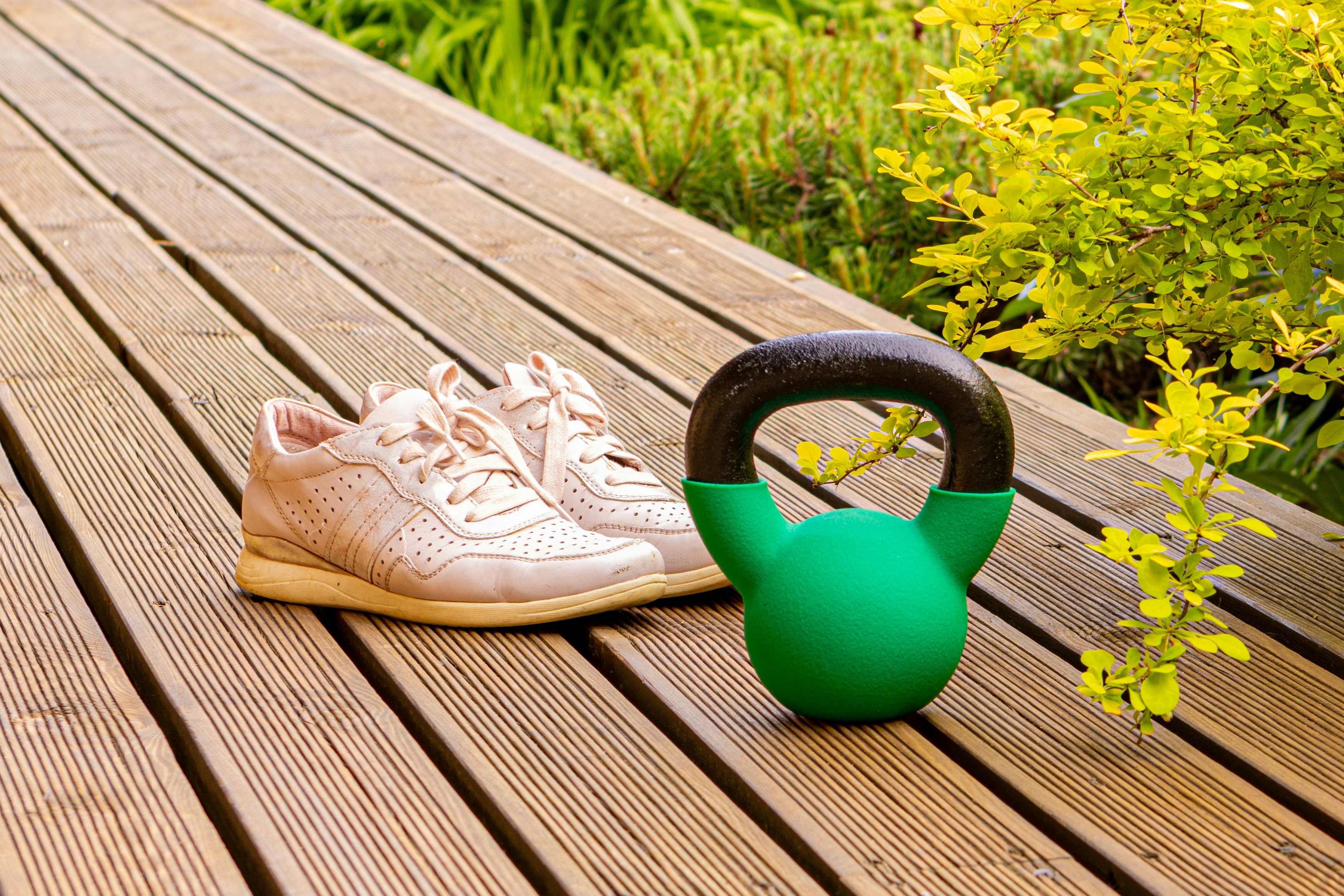 Pale pink trainers and a green kettlebell on a sunlit wooden deck beside leafy shrubs.