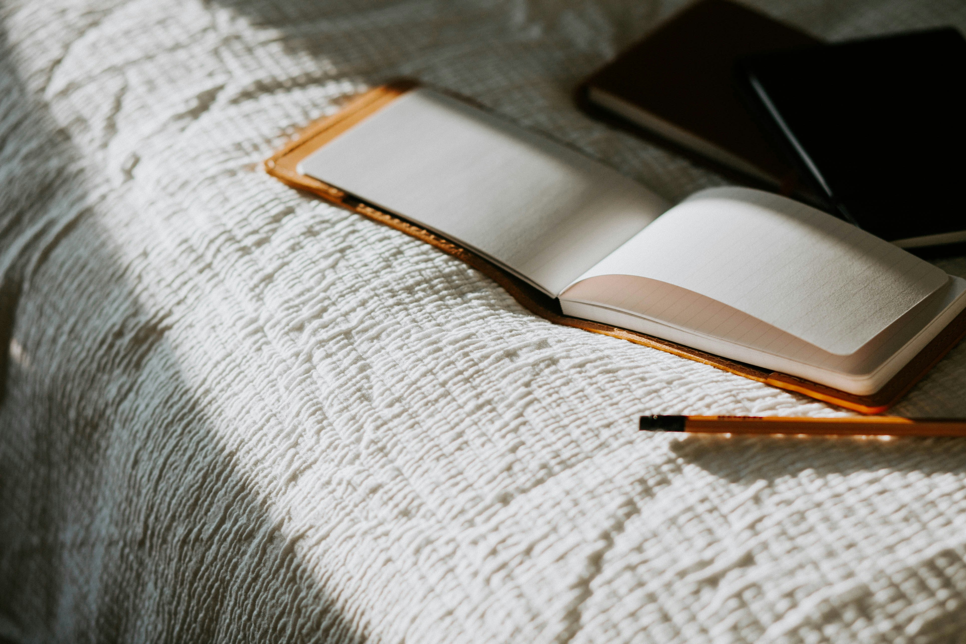 An open blank notebook and pencil on a white quilted bedspread, with a dark notebook nearby.